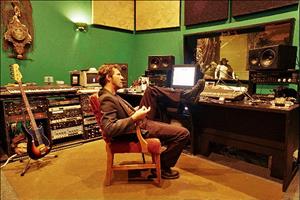 Owner Gary Mula sits in an office chair in the control room of The Dutchman rehearsing and recording studio, his left foot resting on the desk that holds the mixing board and other recording equipment. An ornate clock hangs on the wall to the rear, and a musician can be seen in the window that open into the recording room. Beneath that is more recording equipment, a guitar, and an electric keyboard.